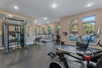 A spacious gym with a variety of exercise equipment and a view of the outdoors through large windows. at Bridge at Heritage Woods, Austin
